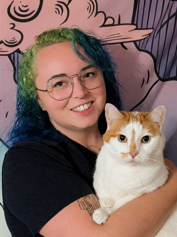 A smiling person with green and blue hair and round glasses holds a white and orange cat in front of a pink backdrop.