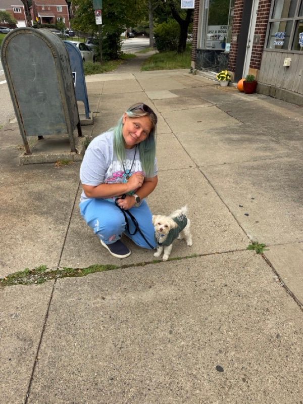 A smiling person with green-tinted hair crouches on a sidewalk beside a small fluffy white dog wearing a green sweater.