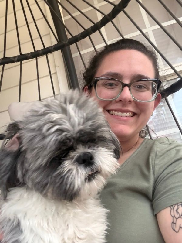 A smiling person with glasses and a green shirt holds a gray and white fluffy dog while sitting in a wicker chair.
