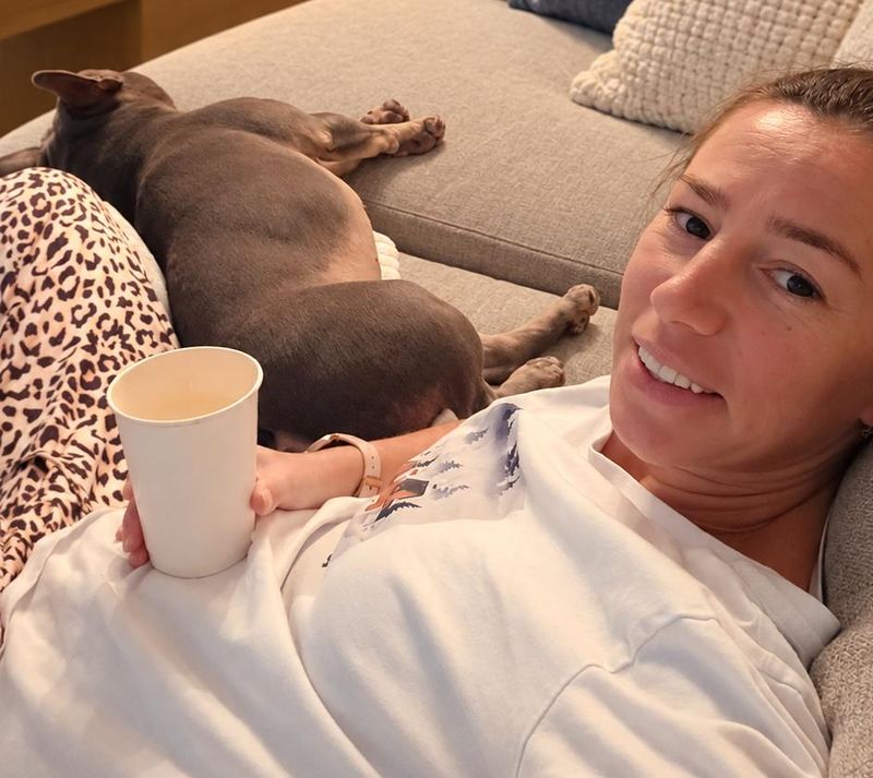 A smiling woman lies on a gray couch holding a white paper cup, with a gray dog stretched out sleeping beside her.