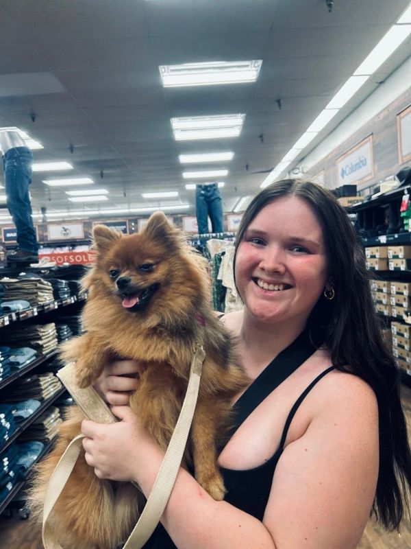 A smiling woman with long dark hair holds a fluffy orange dog with a tan leash in a store aisle with shelves of folded jeans.
