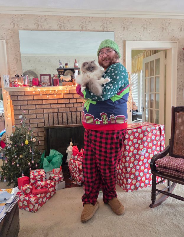 A smiling person in a green knit hat and glasses holds a fluffy cat in a holiday living room with wrapped gifts.