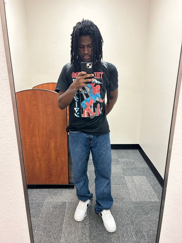 A person takes a mirror selfie in an office hallway, wearing a black graphic t-shirt, blue jeans, and white sneakers.