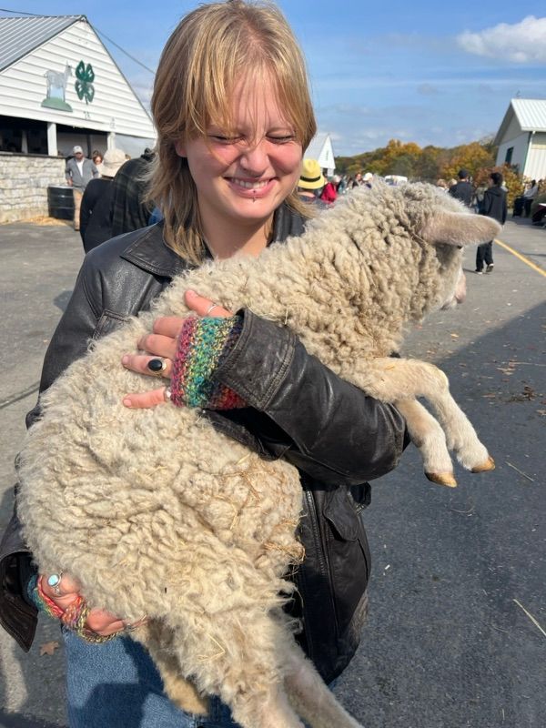 A smiling person in a black leather jacket and colorful knit wrist warmers holds a woolly white lamb outdoors on a paved lot.
