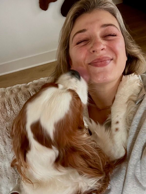 A smiling woman with eyes closed holds a brown and white dog that is nuzzling against her face.