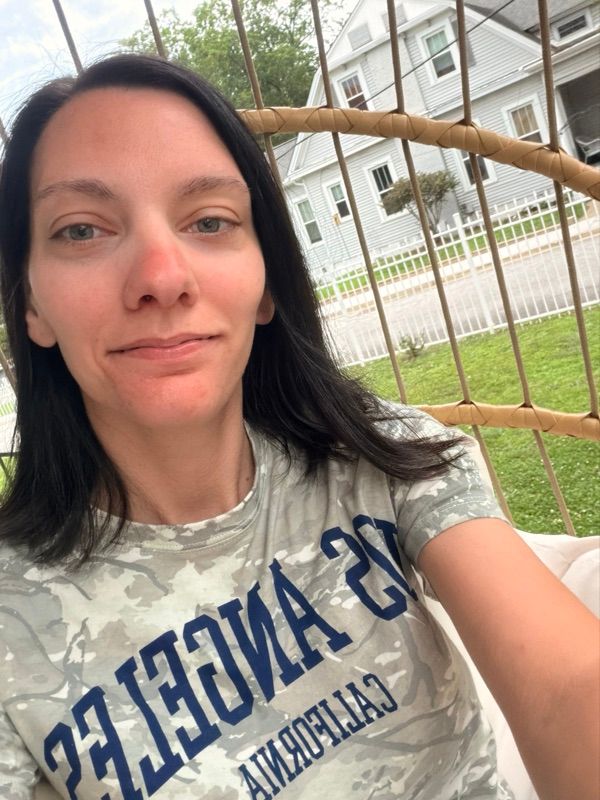 A woman with shoulder-length dark hair takes a selfie outdoors in a camouflage shirt, with a fence and lawn behind her.