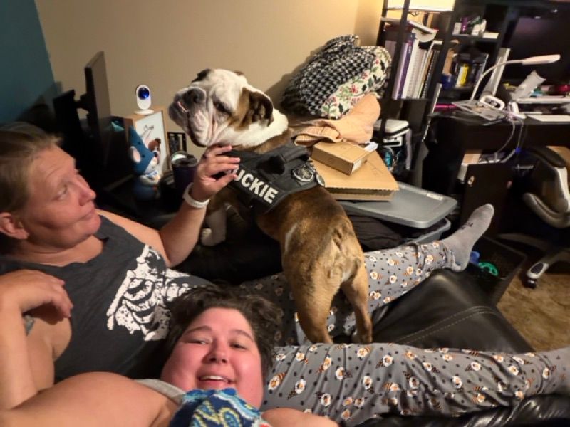 Two people lie on a black couch in a cluttered room while a brown and white bulldog wearing a black vest stands between them.